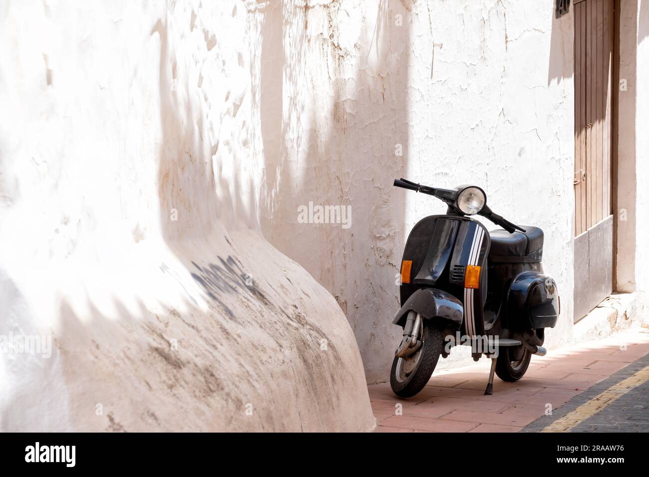 Ein schwarzer vespa-Motorroller parkt auf dem Bürgersteig in der Altstadt von Estepona, Spanien. Der Roller befindet sich neben einer alten, weiß gewaschenen Wand Stockfoto