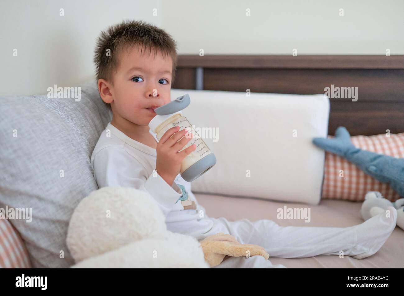 2 Jahre alter Junge sitzt auf dem Bett und trinkt fröhlich Milch aus einer mit Stroh ausgestatteten Flasche, umgeben von Spielzeugfreunden am Morgen in Pyjamas Stockfoto