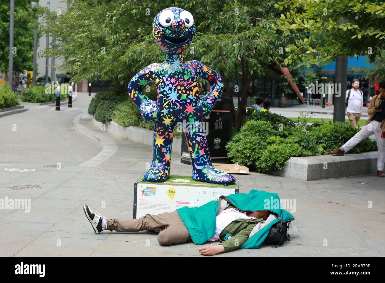 London, Großbritannien. 02. Juli 2023 Ein Obdachloser schläft bei der Skulptur von Morph, der Figur der Kinderserie, am Aldgate Square. Die menschengroße Statue wurde zusammen mit der Kinderrollstuhlorganisation Whizz-Kidz geschaffen. Kredit: Waldemar Sikora/Alamy Live News Stockfoto