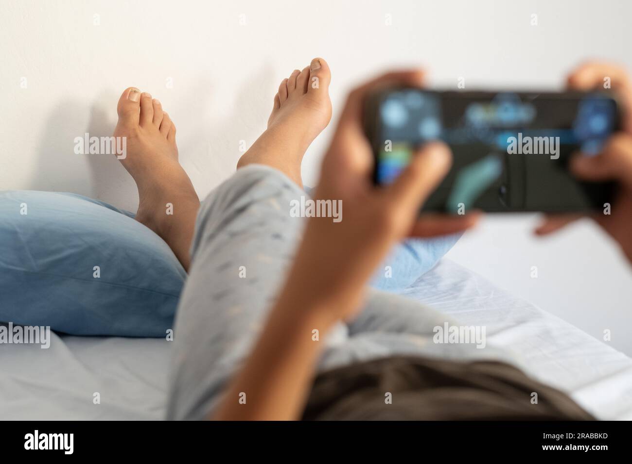 Barfuß Teenager Junge mit gekreuzten Beinen ruht auf dem Kissen mit Handy auf dem Bett. Ein Teenager, der morgens Videospiele in seinem Zimmer spielt. Stockfoto