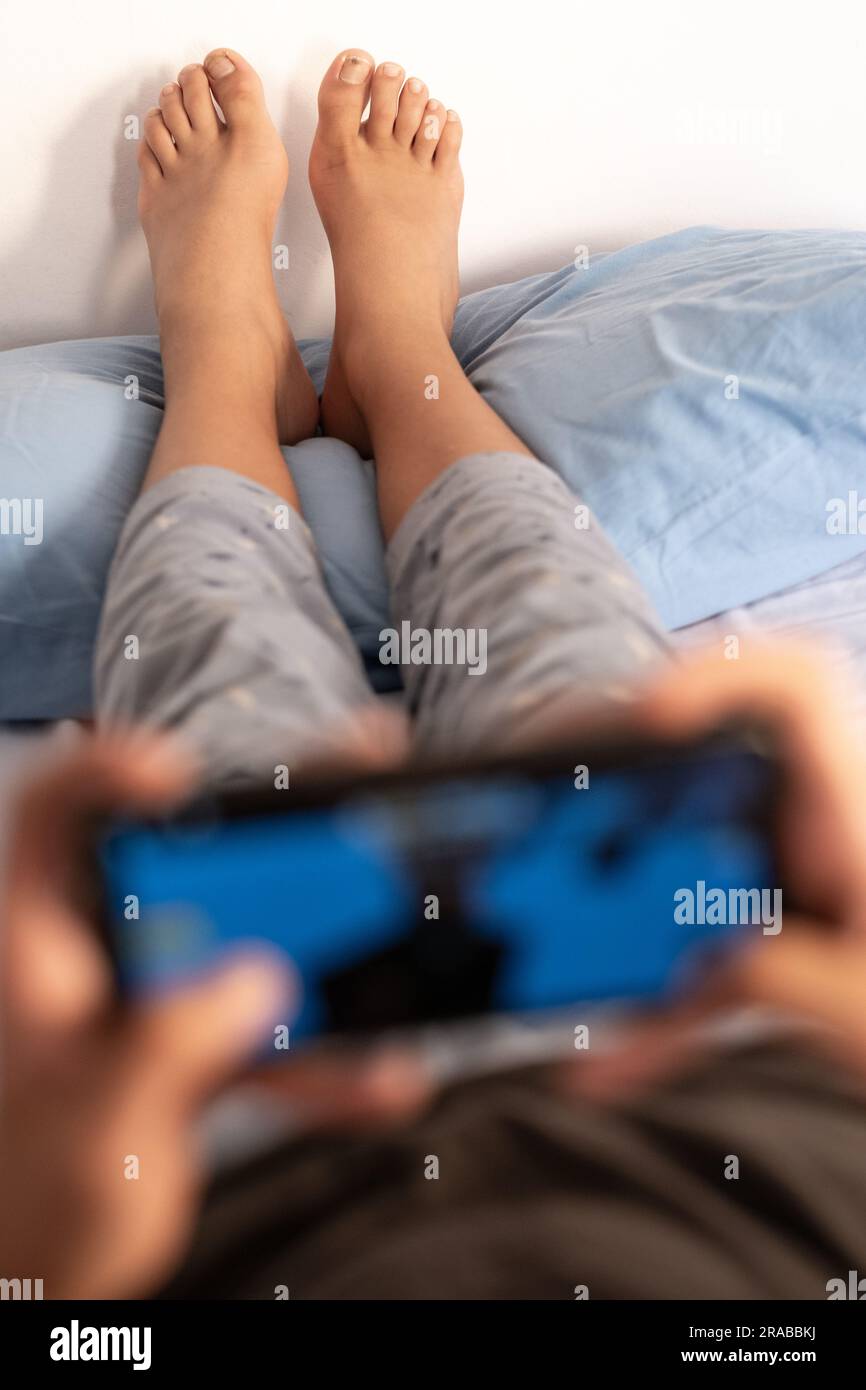 Ein Teenager im Schlafanzug am Morgen, der auf dem Bett liegt, spielt Videospiele, während seine Füße auf dem Kissen ruhen. Fokus auf Hintergrund und Kopierbereich. Stockfoto