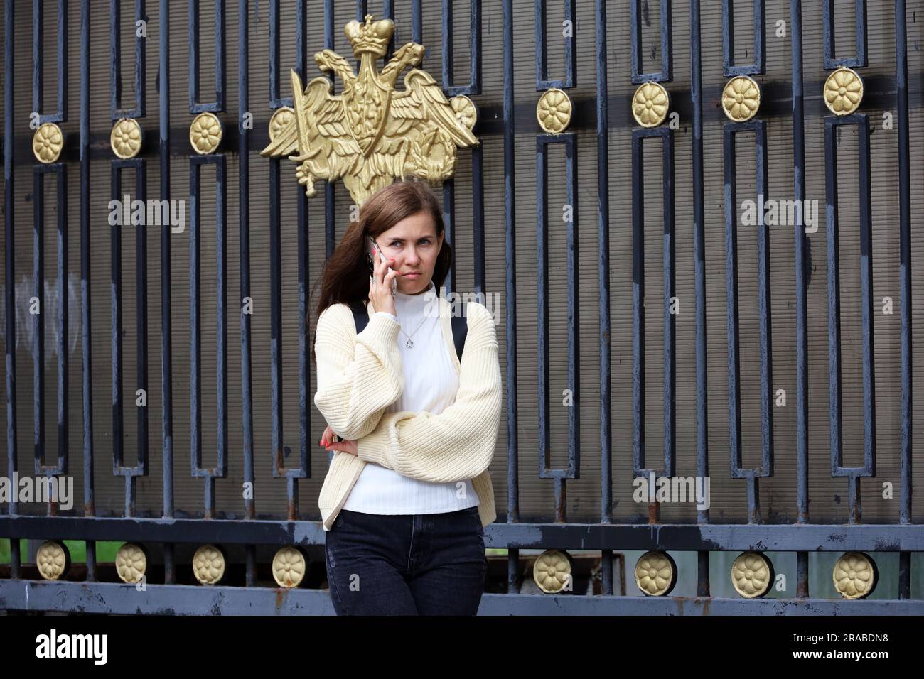 Eine Frau, die auf der Straße am Zaun mit einem Handy spricht, mit einem offiziellen Emblem des russischen Verteidigungsministeriums Stockfoto