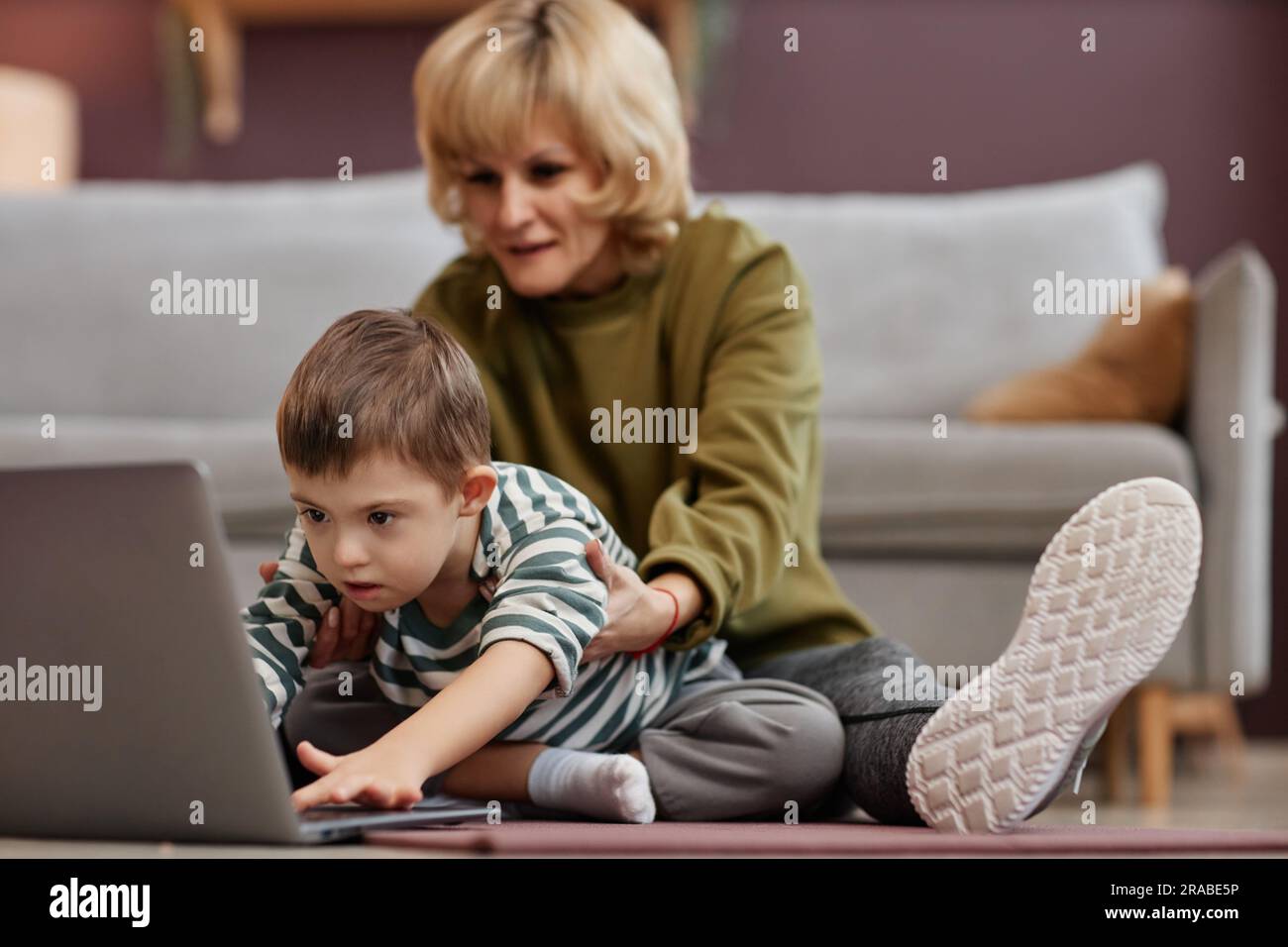 Porträt eines kleinen Jungen mit Down-Syndrom, der zu Hause ein Notebook benutzt und eine fürsorgliche Mutter als Assistentin hat Stockfoto