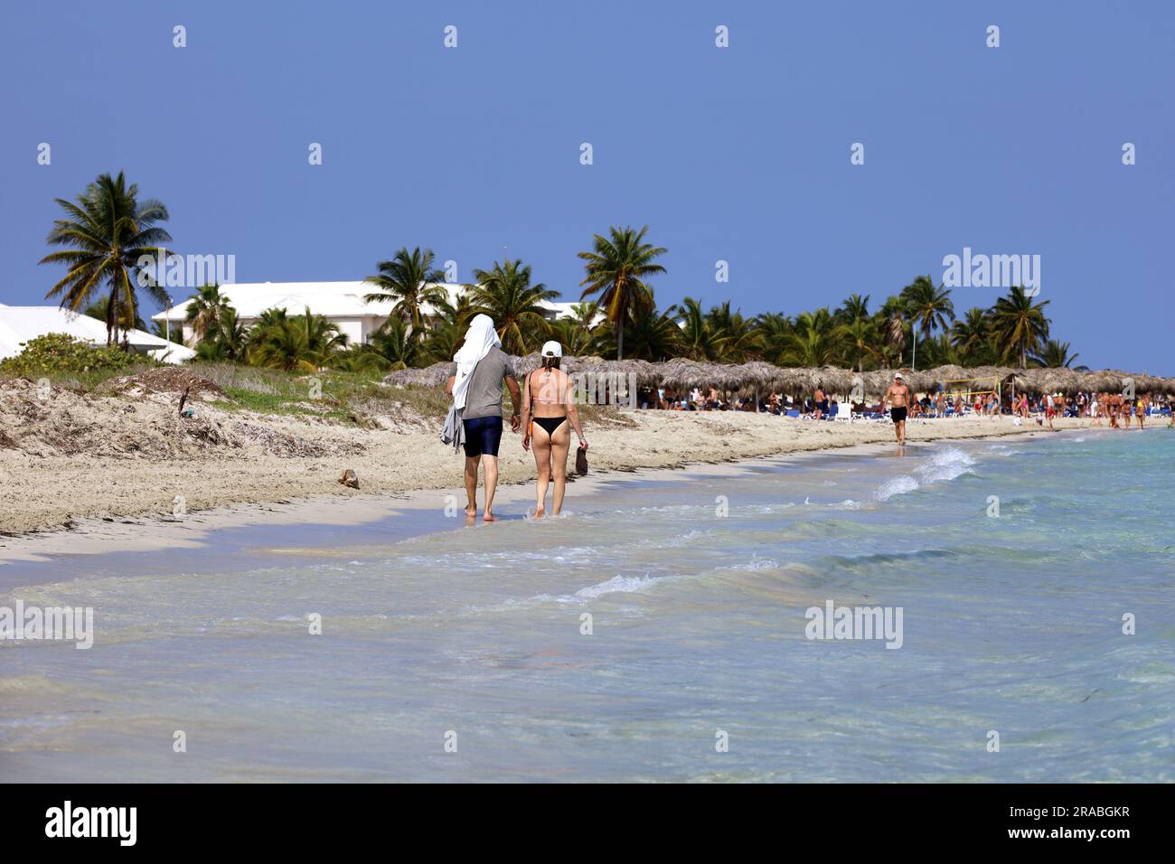 Ein Paar in Badebekleidung, das am Sandstrand entlang der Meereswellen spaziert. Tropisches Resort mit Kokospalmen und Korbschirmen Stockfoto