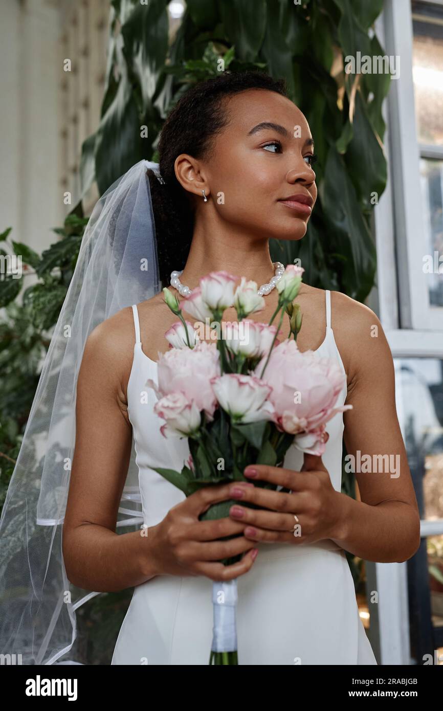 Vertikales Porträt einer schönen schwarzen Frau als junge Braut, die ein einfaches Hochzeitskleid trägt und einen Bouquet zur Seite hält Stockfoto
