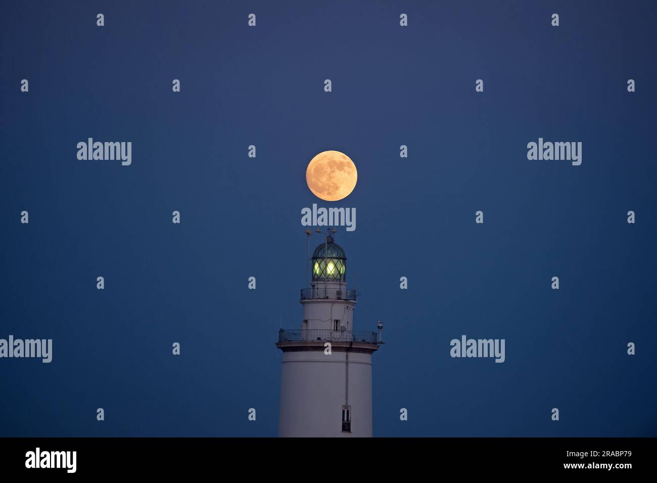 Der Buckmond, der am Himmel über einem Leuchtturm aufgeht, ist im Hafen von Malaga zu sehen. Anfang Juli findet der Vollmond der Hirsche als eines der beeindruckendsten astronomischen Ereignisse des Monats statt. Wenn der Vollmond stattfindet, erscheint er größer und heller als normal. Stockfoto