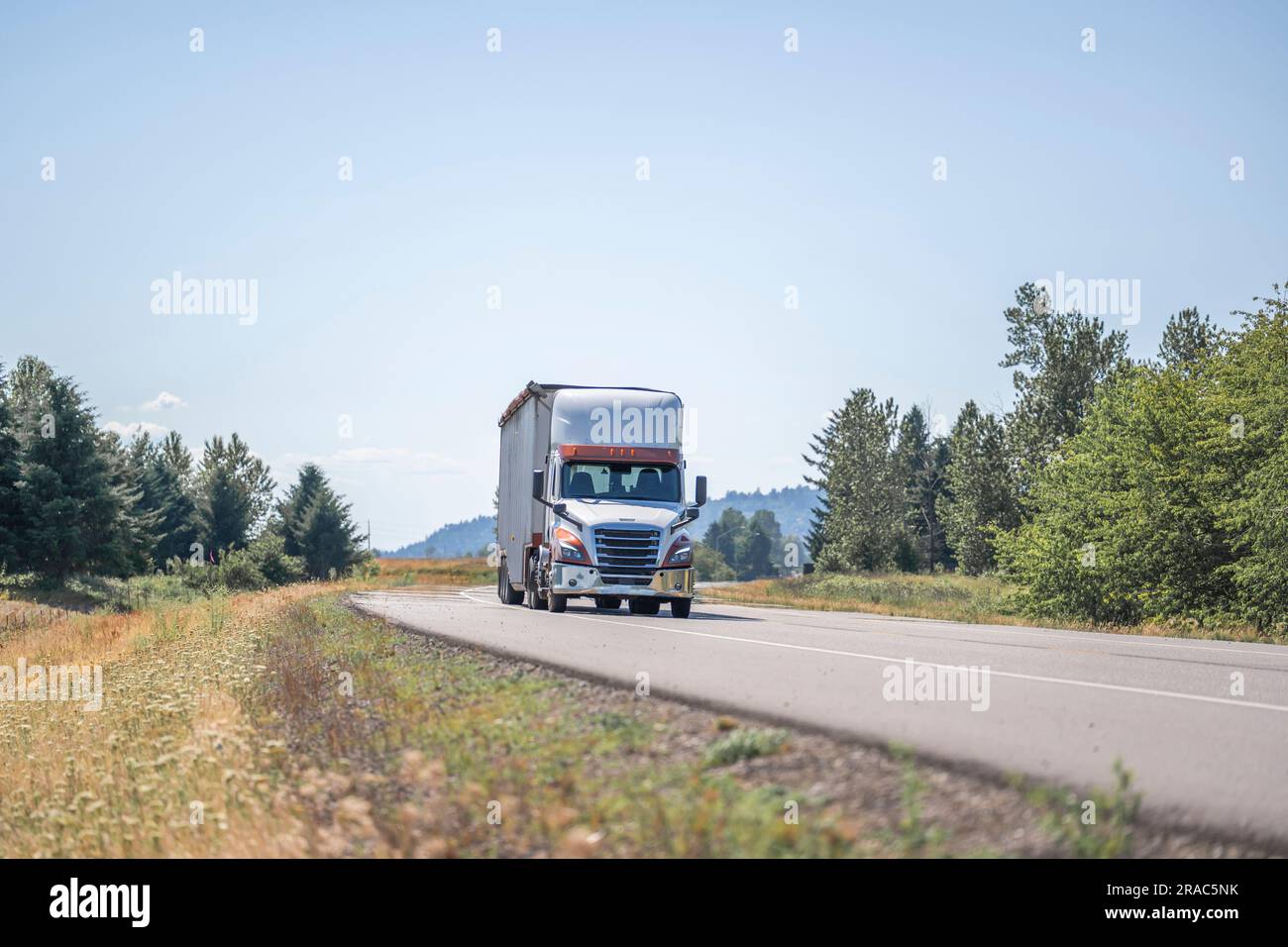 Industrielles weißes Day Cab großer Truck mit Motorhaube klassischer Sattelschlepper, der gewerbliche Fracht in einem abgedeckten Sattelanhänger transportiert, während der schmale Sommer-sce gefahren wird Stockfoto