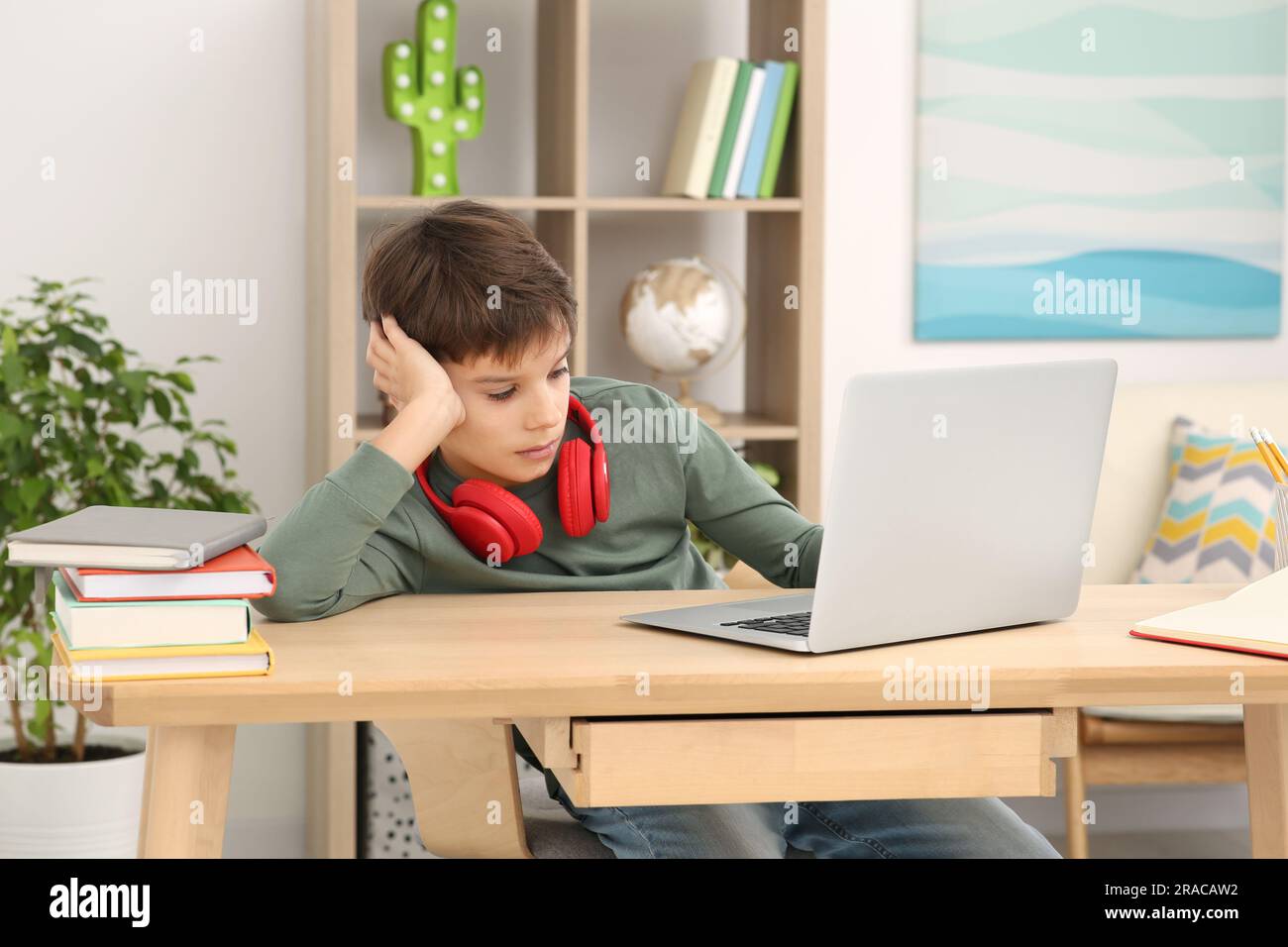 Müder Junge mit roten Kopfhörern, der am Schreibtisch im Zimmer ein Notebook benutzt. Zu Hause Stockfoto