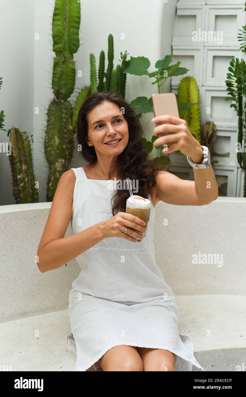 Porträt einer 40-jährigen russin, die sich nicht schminkt, mit einem Glas Eiskaffee in weißem Kleid sitzt und ein Selfie macht Stockfoto