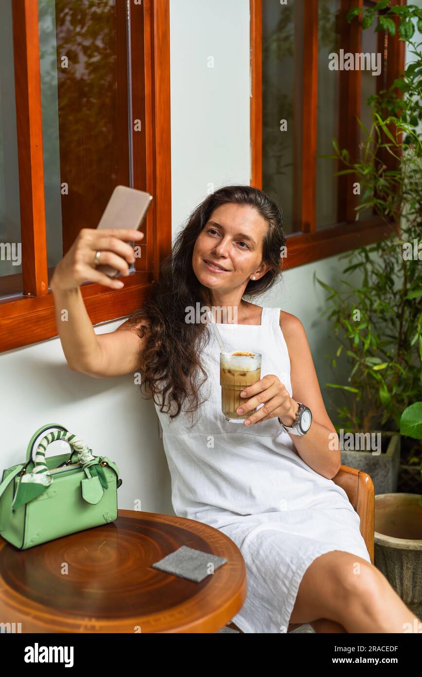 Porträt einer 40-jährigen russin, die sich nicht schminkt, mit einem Glas Eiskaffee in weißem Kleid sitzt und ein Selfie macht Stockfoto