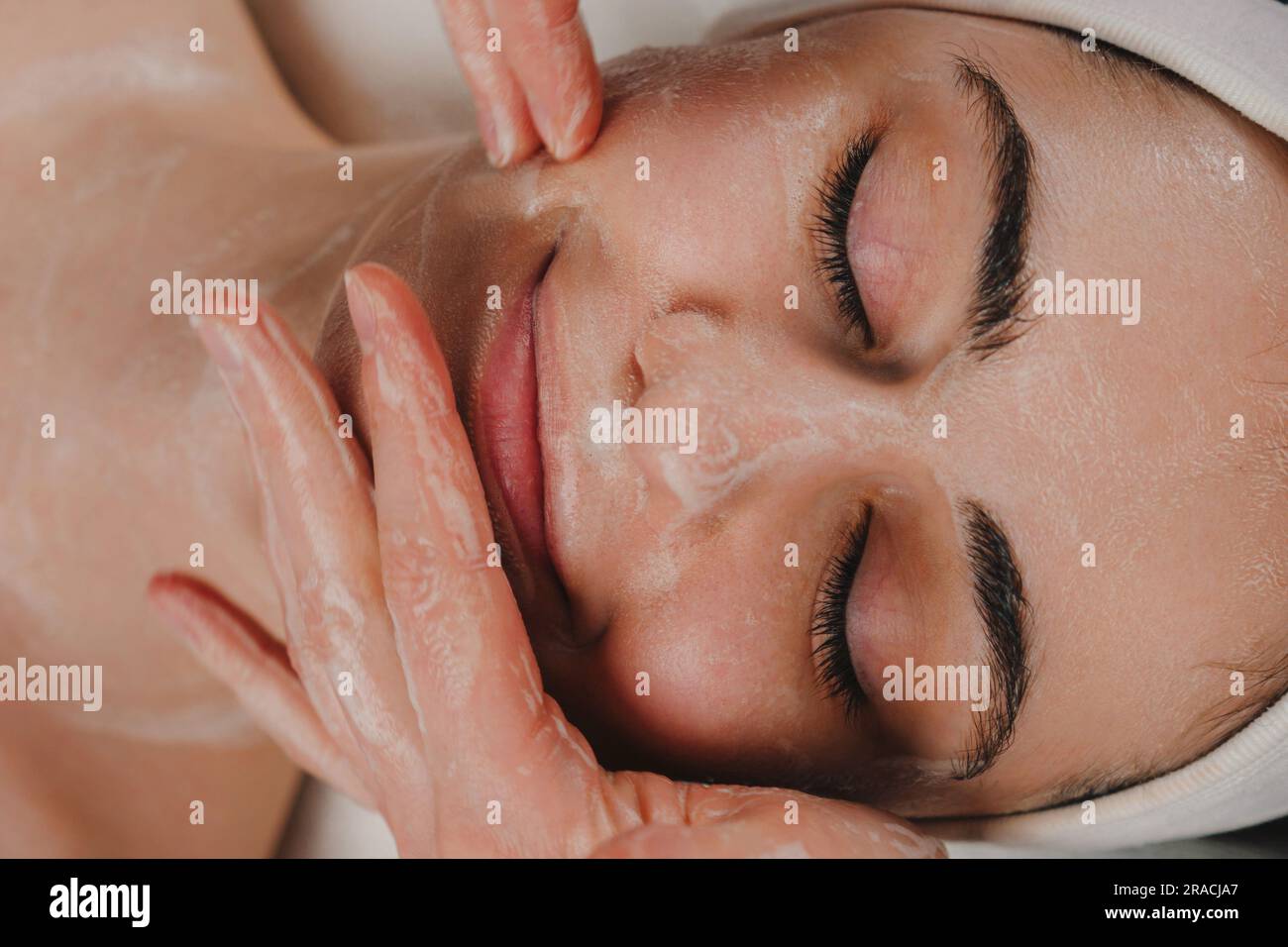 Crop anonyme Frau mit Gesichtsmaske auf Gesicht einer jungen Frau mit geschlossenen Augen in einem Spa-Salon. Stockfoto