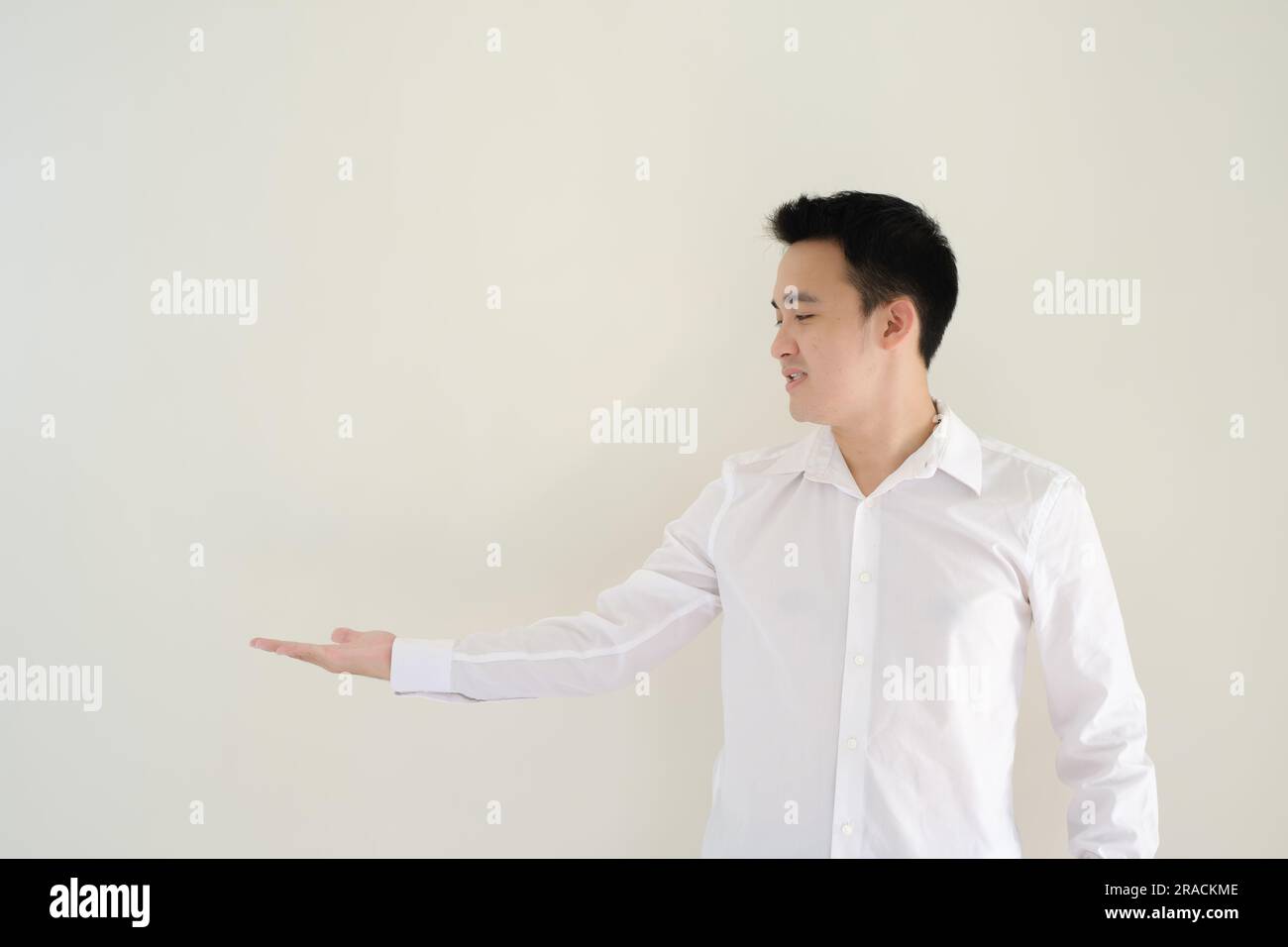Ein junger asiatischer Mann, der ein weißes Hemd trägt, öffnet seine rechte Hand zur rechten Seite und sieht sie an. Isolierter weißer Hintergrund. Stockfoto Ein junger asiatischer Mann, der ein weißes Hemd trägt, öffnet seine rechte Hand zur rechten Seite und sieht sie an. Isolierter weißer Hintergrund. Stockfoto