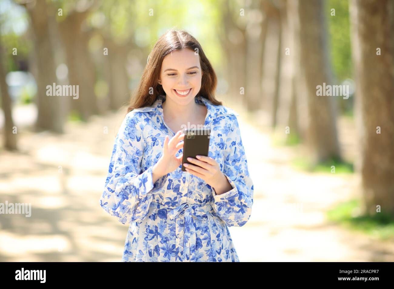 Porträt einer glücklichen Frau, die mit dem Handy in einem sonnigen Park spaziert Stockfoto