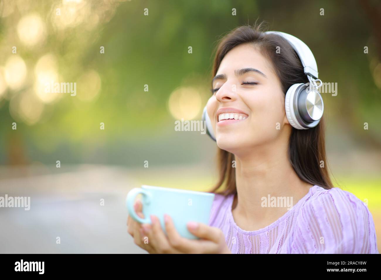 Glückliche Frau mit Kopfhörern, die Musik hört, die Kaffeetasse in einem Park hält und frische Luft atmet Stockfoto