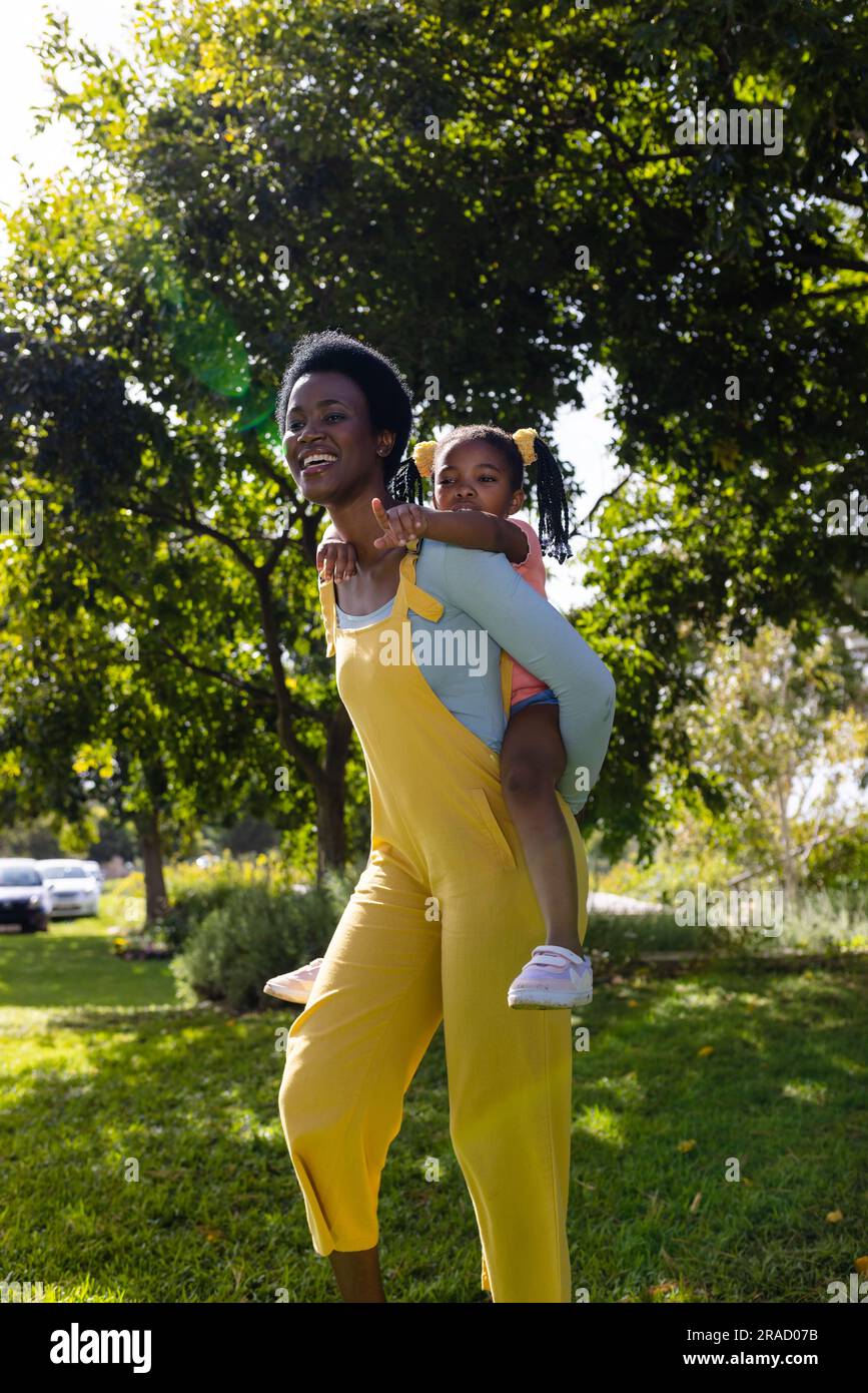 afroamerikanische Mutter, die sich mit dem Huckepack beschäftigt, fröhliche Tochter, während sie auf dem grasbewachsenen Feld im Park spaziert Stockfoto