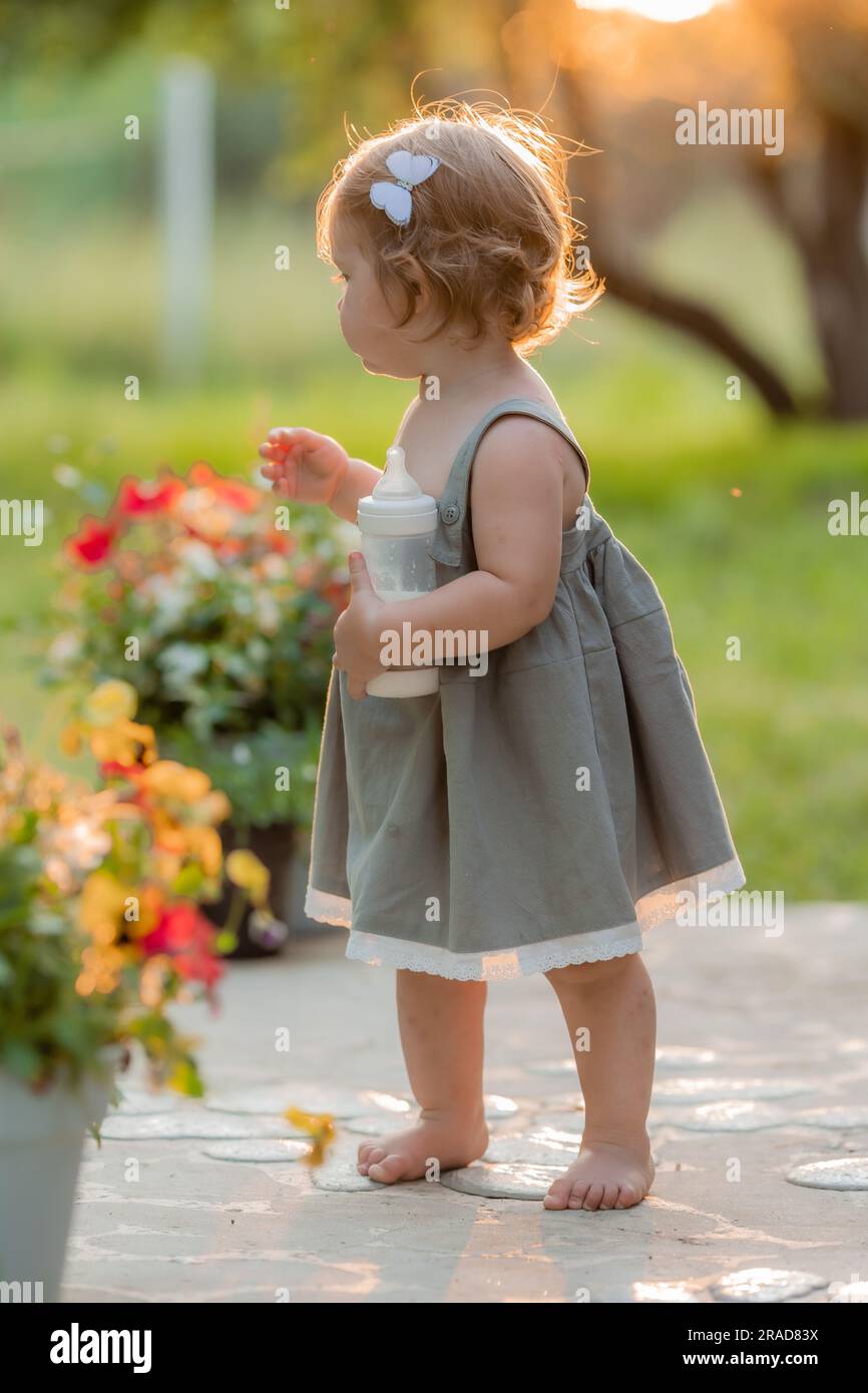 Ein kleines Mädchen in einem Sommerkleid aus Baumwolle geht im Sommer bei Sonnenuntergang im Park spazieren und trinkt Milch aus einer Babyflasche. Babynahrung. Hochwertiges Foto Stockfoto