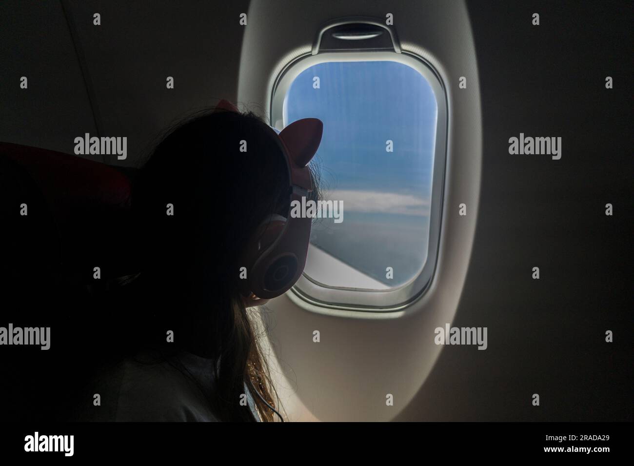 Ein Mädchen, das den Himmel aus dem Fenster eines Flugzeugs beobachtet. Sie hat Kopfhörer auf dem Kopf. Stockfoto
