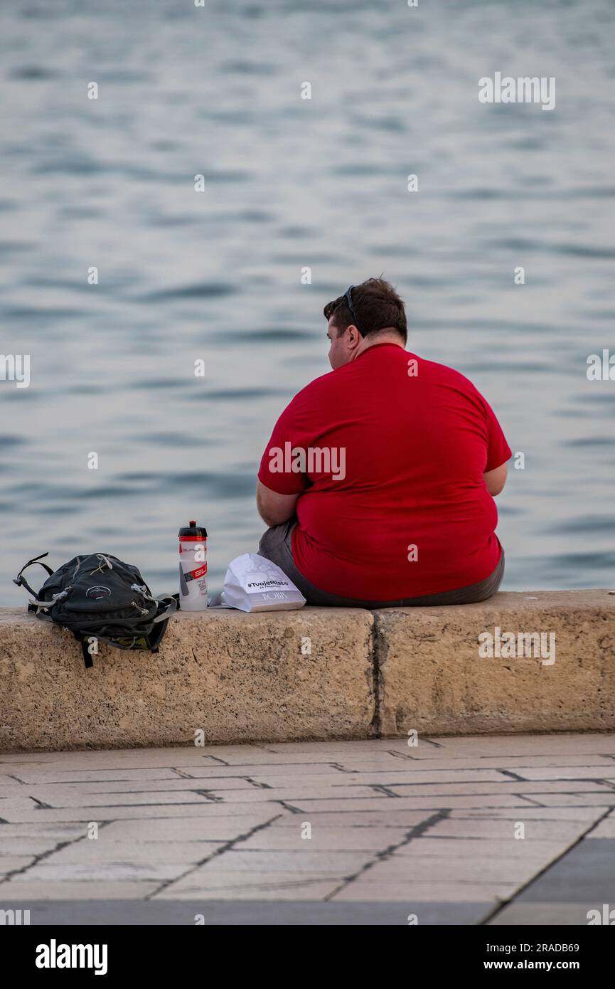 Übergewichtiger Mann sitzt an einer Wand am Meer, übergewichtiger Mann sitzt an einer Wand in der Nähe des Meeres, großer übergewichtiger junger Mann sitzt mit Blick aufs Meer Stockfoto