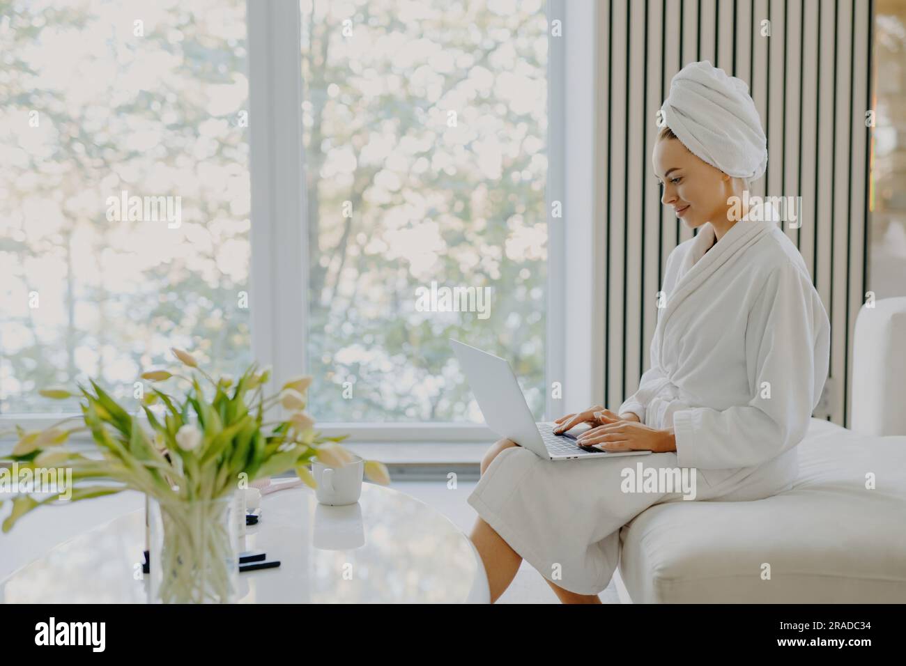 Erfrischte Frau im Bademantel, Handtuch am Kopf, arbeitet am Laptop, sitzt auf dem Sofa, posiert am Fenster in einem gemütlichen Zimmer. Stockfoto