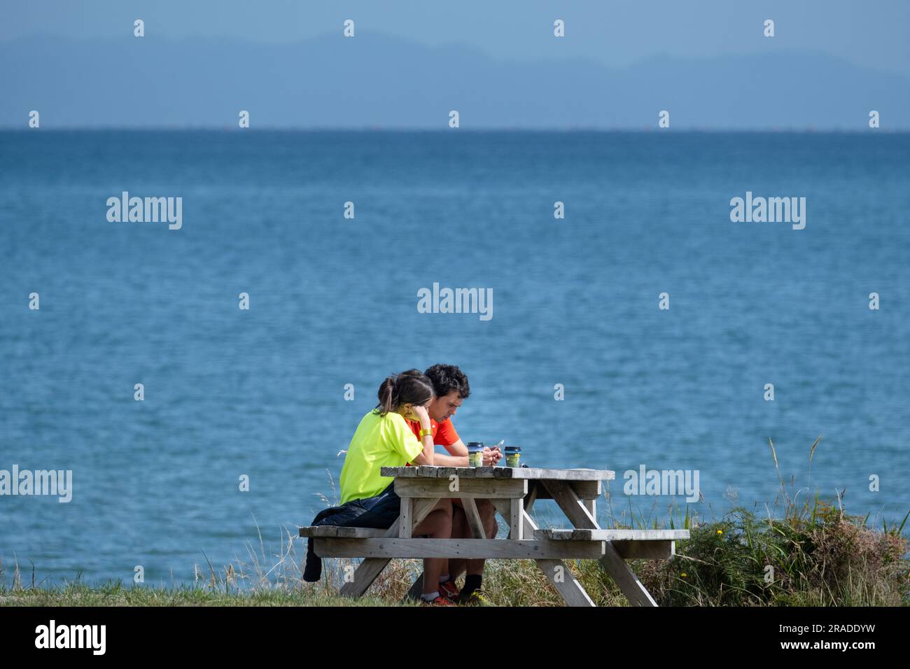 Ein Paar mit Handy und Kaffee am Strand Pōhara, Takaka Küstenregion in Golden Bay, Abel Tasman Nationalpark auf Südinsel, Neuseeland. Stockfoto