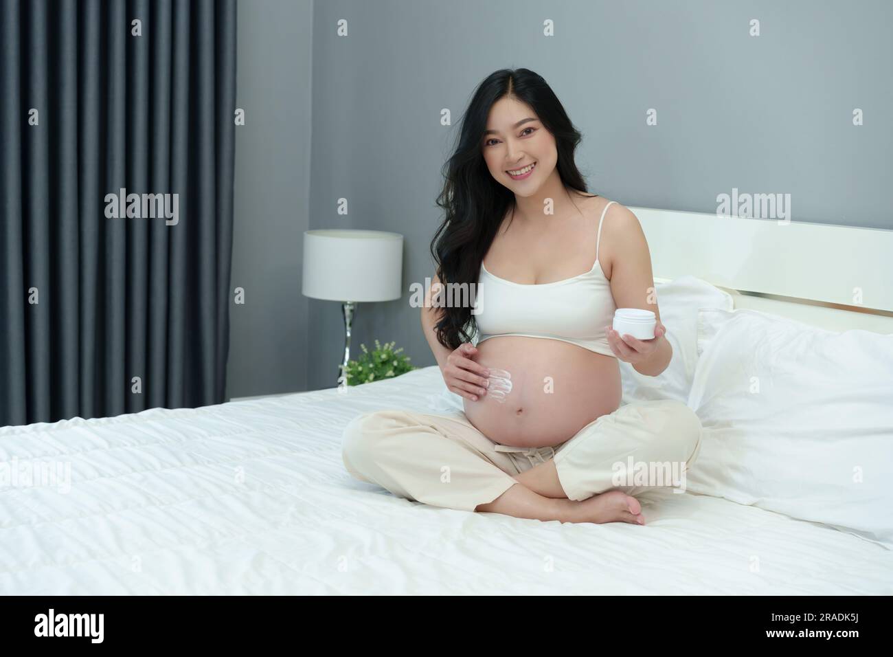 Schwangere Frau streichelt auf dem Bett Creme auf den Bauch, um die Haut mit Feuchtigkeit zu versorgen Stockfoto