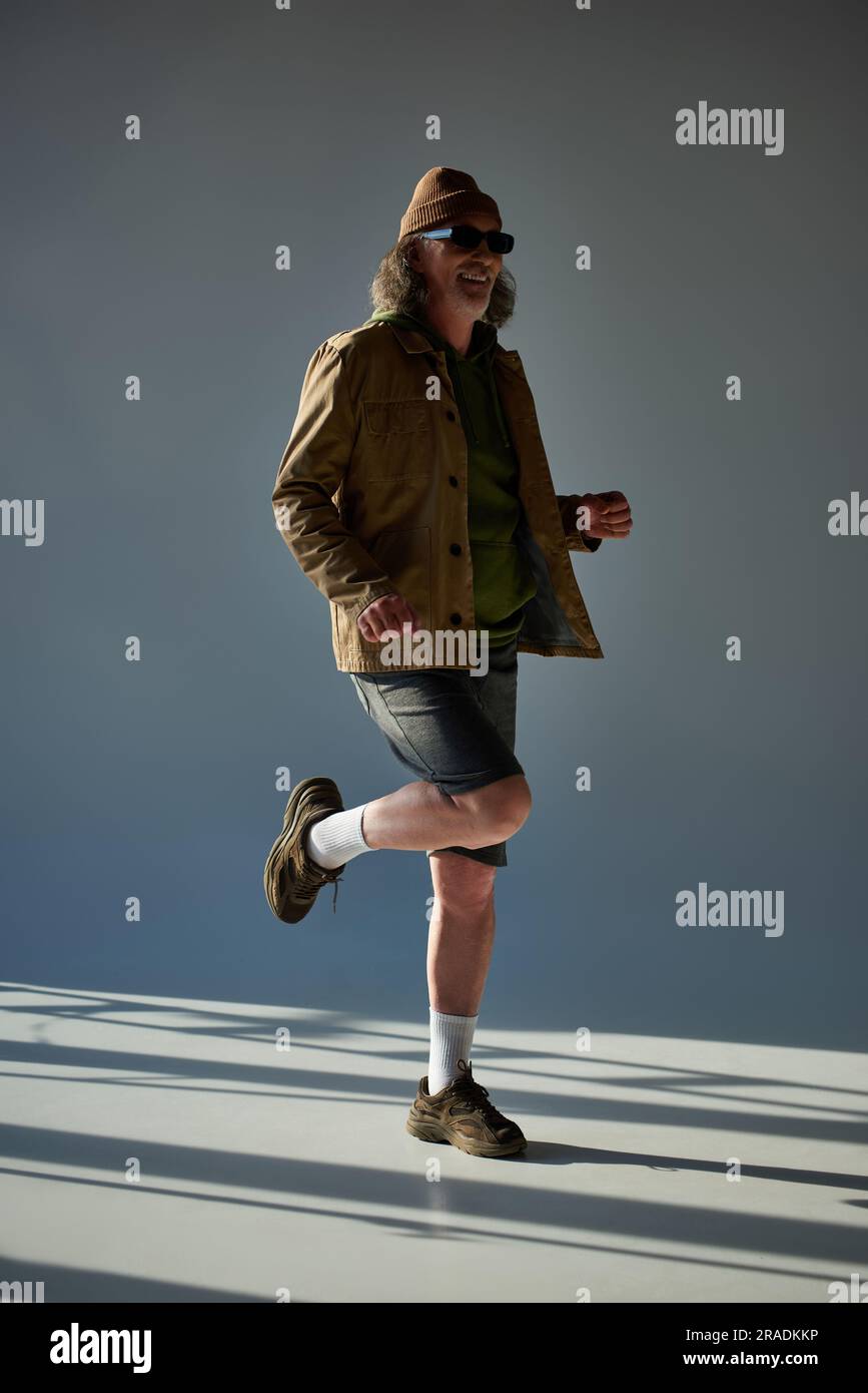 Ein langes, lächelndes und stilvolles männliches Modell, das auf einem Bein auf grauem Hintergrund posiert, mit Beleuchtung, Hipster-Mode, dunkler Sonnenbrille, Jacke und Stockfoto