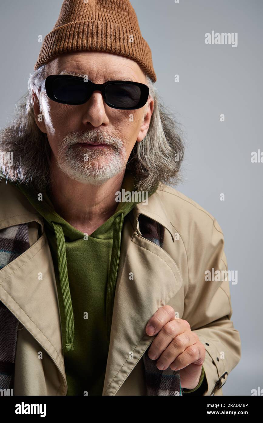 Porträt eines grauhaarigen und bärtigen Mannes mit Mütze, dunkler Sonnenbrille und beigefarbenem Trenchcoat, der auf grauen Hintergrund auf die Kamera blickt, Hipster-Modell Stockfoto
