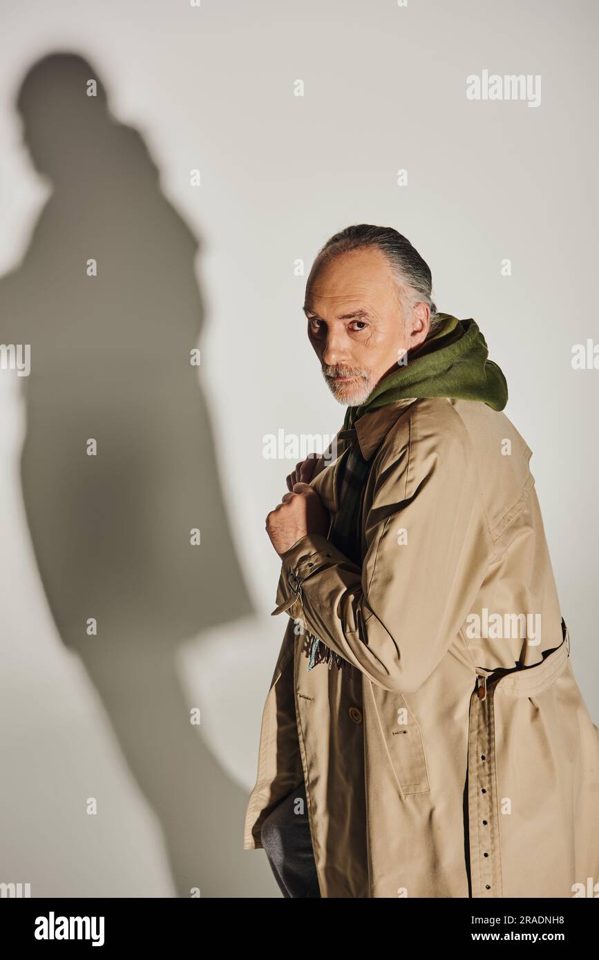 Mode und Alter, persönlicher Stil, selbstbewusster Senior-Mann mit ernstem Gesichtsausdruck, steht in beigefarbenem Trenchcoat und schaut auf die Kamera auf grauem Hintergrund Stockfoto