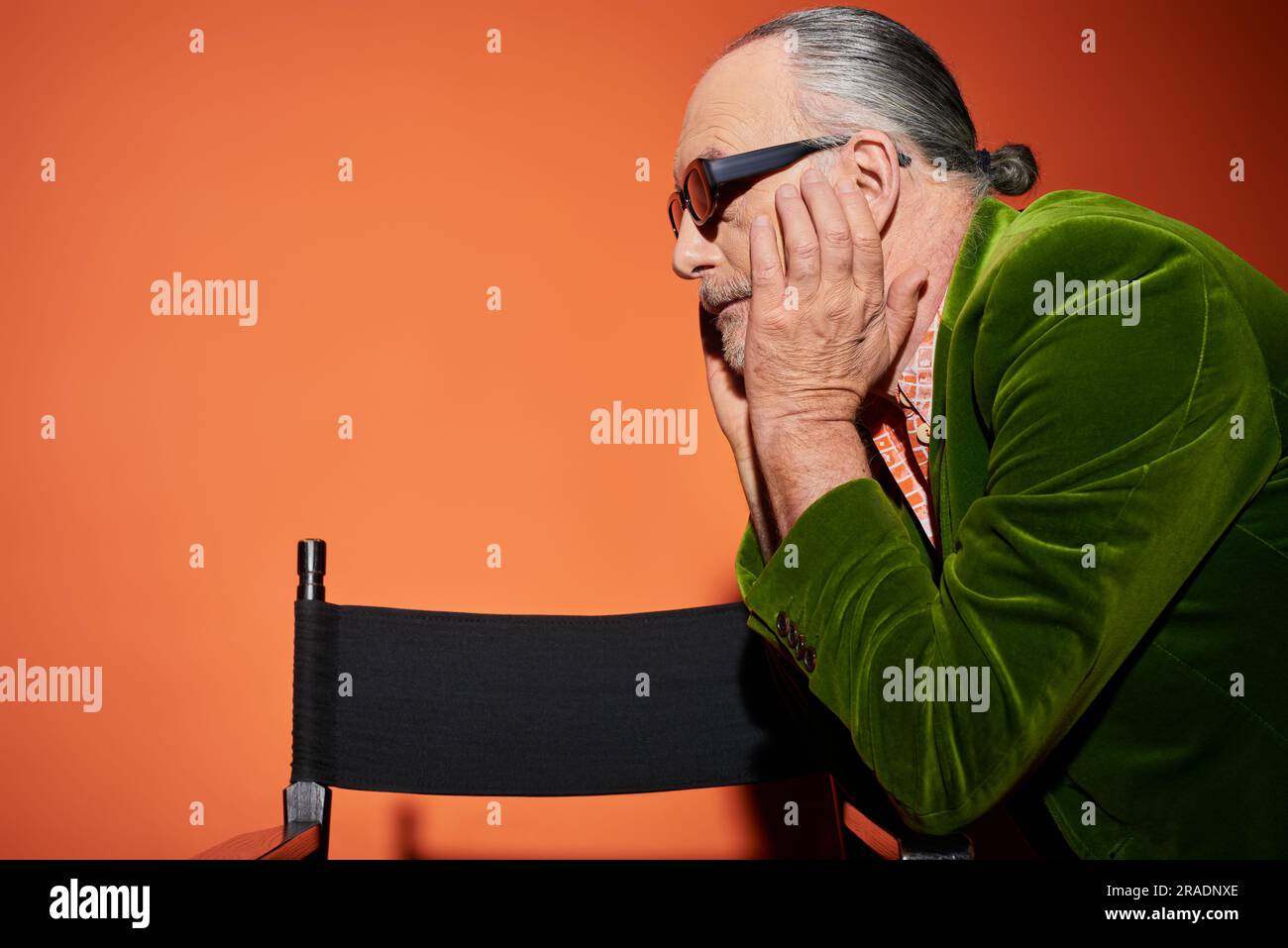Seitenansicht eines älteren, grauhaarigen Modells, das das Gesicht berührt und in die Nähe des Stuhls blickt, mit rotem und orangefarbenem Hintergrund, modischer Look, dunkler Sonnenbrille, grünem Velours Stockfoto