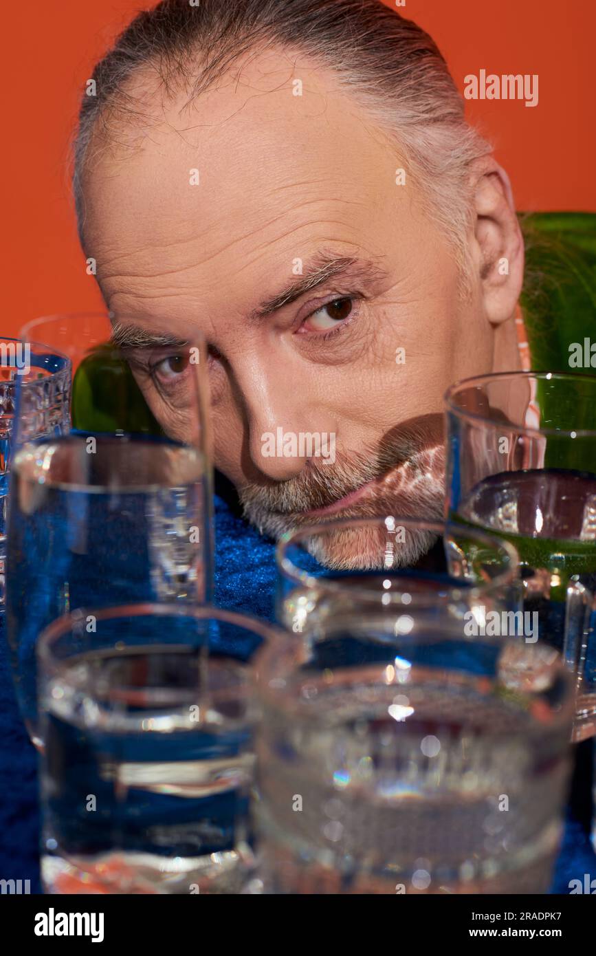 Erinnerungen, Senioren und Nachdenklicher, der in der Nähe von Brillen blickt, klares Wasser auf dem Tisch mit blauem Velours auf orangefarbenem Hintergrund, alternde Bevölkerung, sy Stockfoto