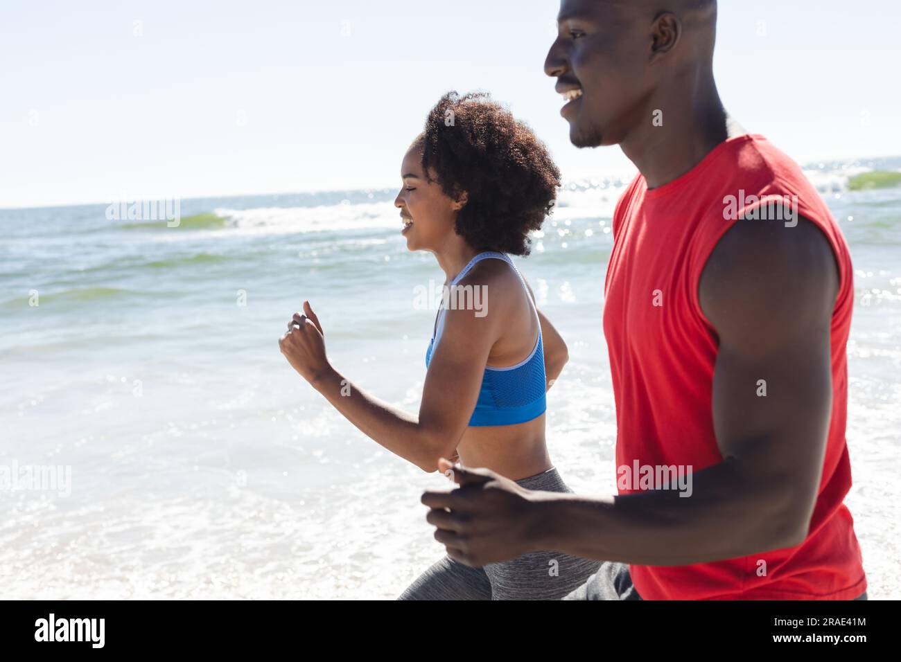 Glückliches, fittes afroamerikanisches Paar, das am sonnigen Strand trainiert, joggt und lächelt Stockfoto