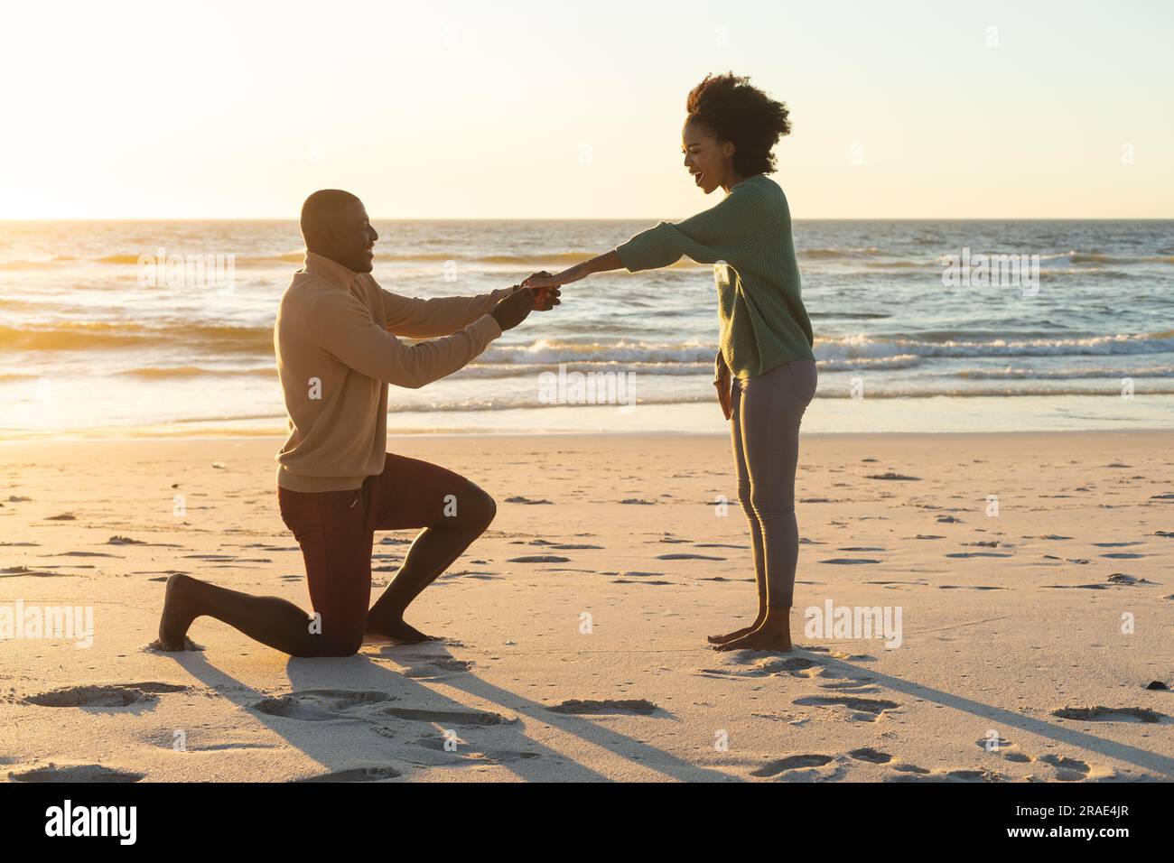 Ein glücklicher afroamerikanischer Mann kniete und machte seiner Freundin am Strand bei Sonnenuntergang einen Heiratsantrag Stockfoto Ein glücklicher afroamerikanischer Mann kniete und machte seiner Freundin am Strand bei Sonnenuntergang einen Heiratsantrag Stockfoto