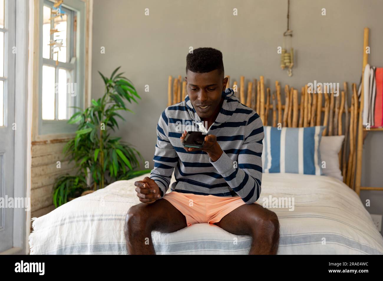 Glücklicher afroamerikanischer Mann, der auf dem Bett sitzt und mit einem Smartphone im sonnigen Ferienhaus am Strand spricht Stockfoto