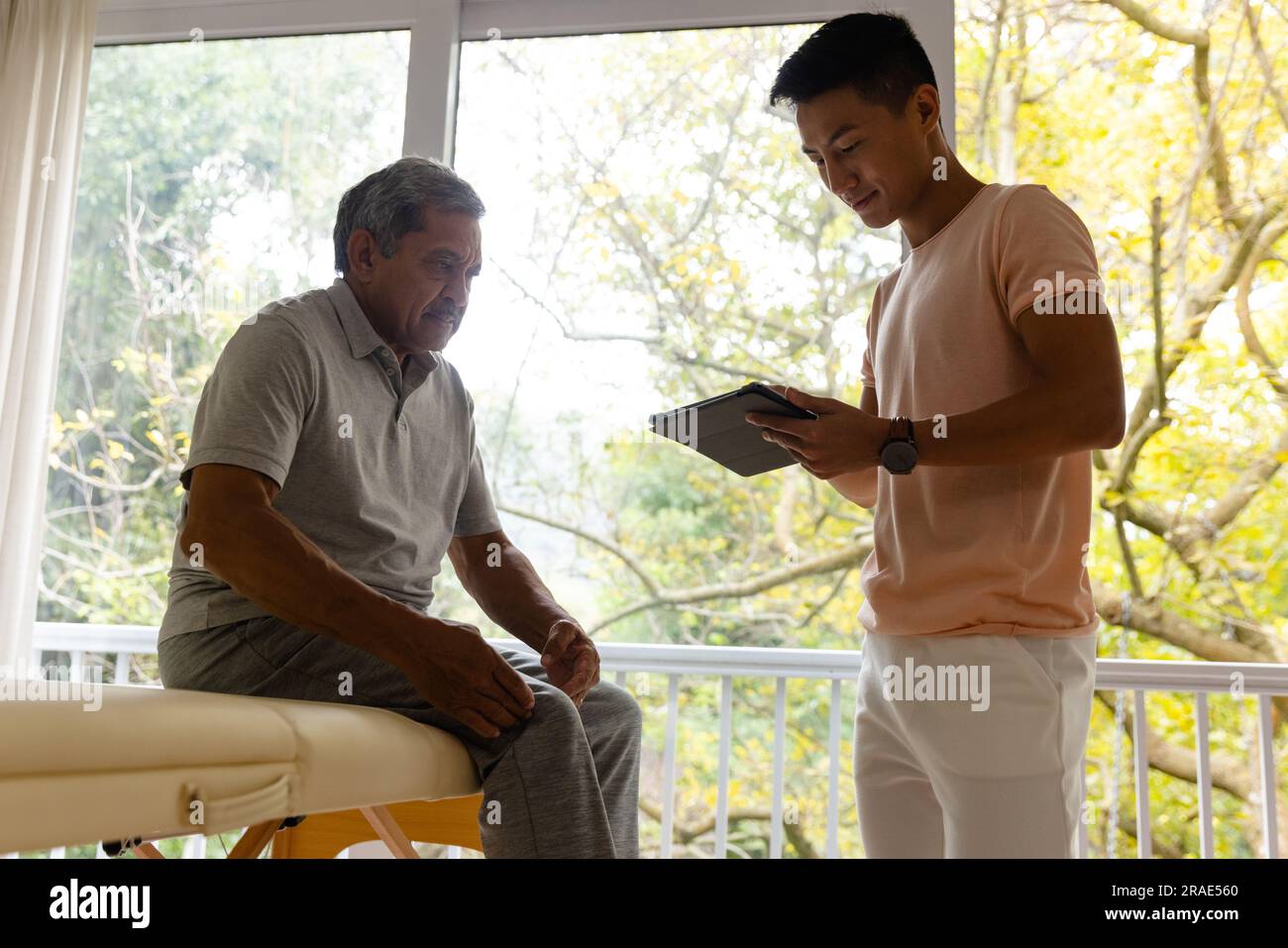 Glücklicher vielseitiger männlicher Physiotherapeut, der ältere männliche Patienten mit Tablet untersucht Stockfoto