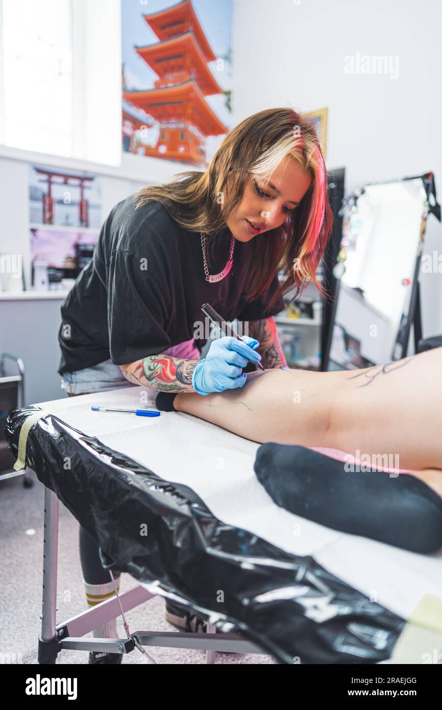 Eine Kundin liegt auf dem Tisch und ein Tattoo-Meister, der mit Freihand-Technik auf ihrem Bein malt. Hochwertiges Foto Stockfoto