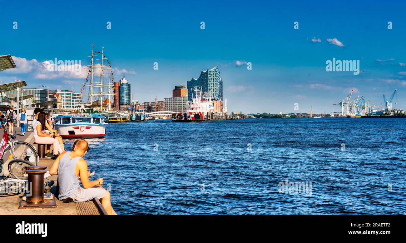 Die Menschen entspannen sich entlang der Uferpromenade am Hamburger Hafen mit der Elbphilharmonie und den Hafenkranichen im Hintergrund Stockfoto