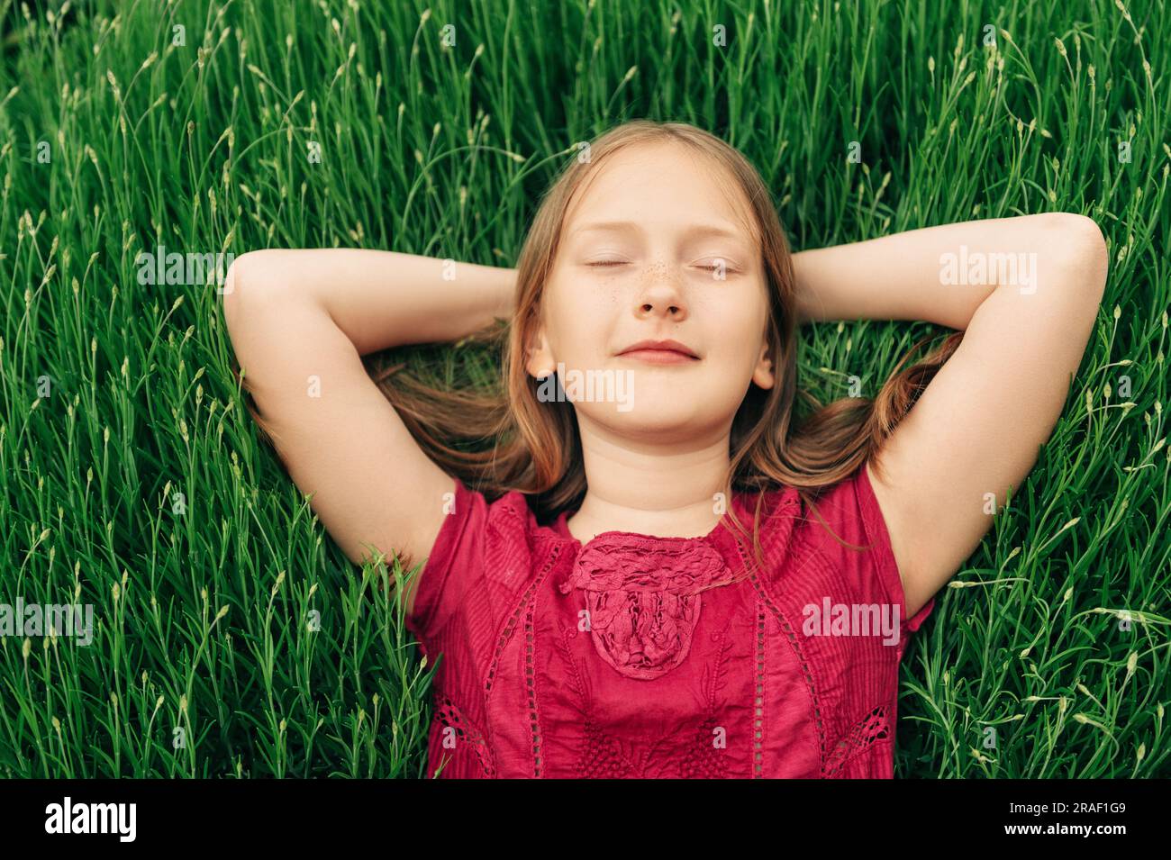 Süßes kleines Mädchen, das auf hellgrünem Gras liegt, die Arme hinter dem Kopf hält, die Augen geschlossen Stockfoto