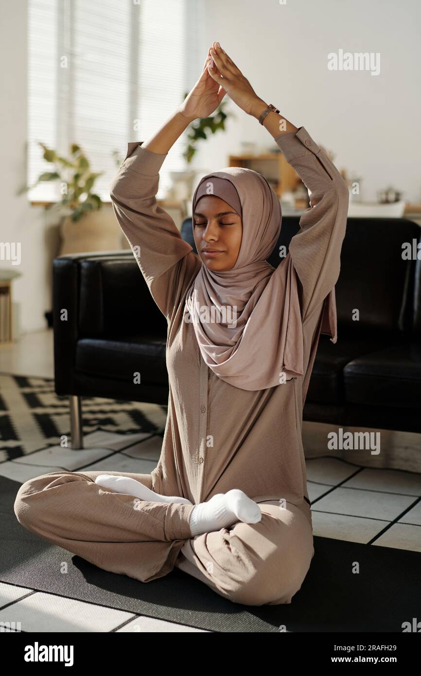 Junge, heitere muslimische Frau in Hijab und Aktivkleidung, die in Lotusposition sitzt, mit zusammengelegten Händen über dem Kopf und geschlossenen Augen Stockfoto