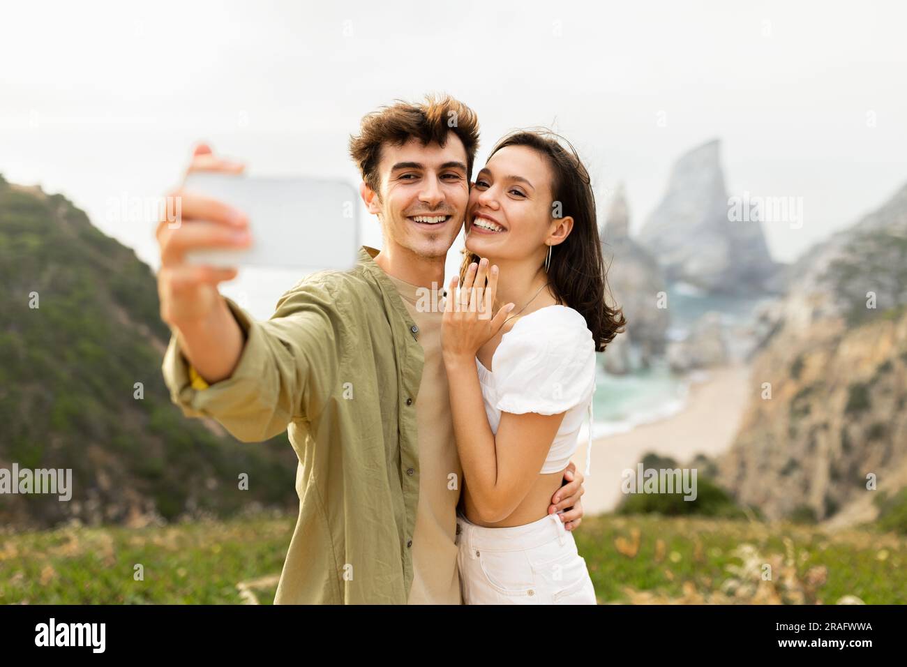 Glückliches europäisches Paar nach der Verlobung, das Selfie auf dem Handy machte, auf einer Felsklippe in der Nähe des Ozeans stand Stockfoto