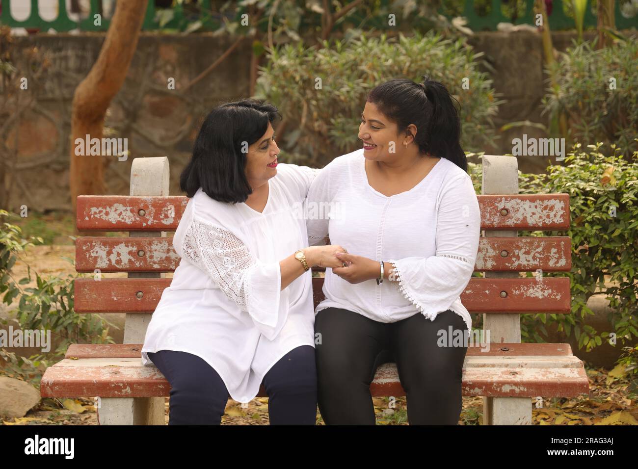 Glückliche indische Mutter und Tochter in weißem Kleid, die wunderschöne Momente auf der Bank im Stadtpark haben. Liebende Mutter und liebenswerte Tochter. Stockfoto