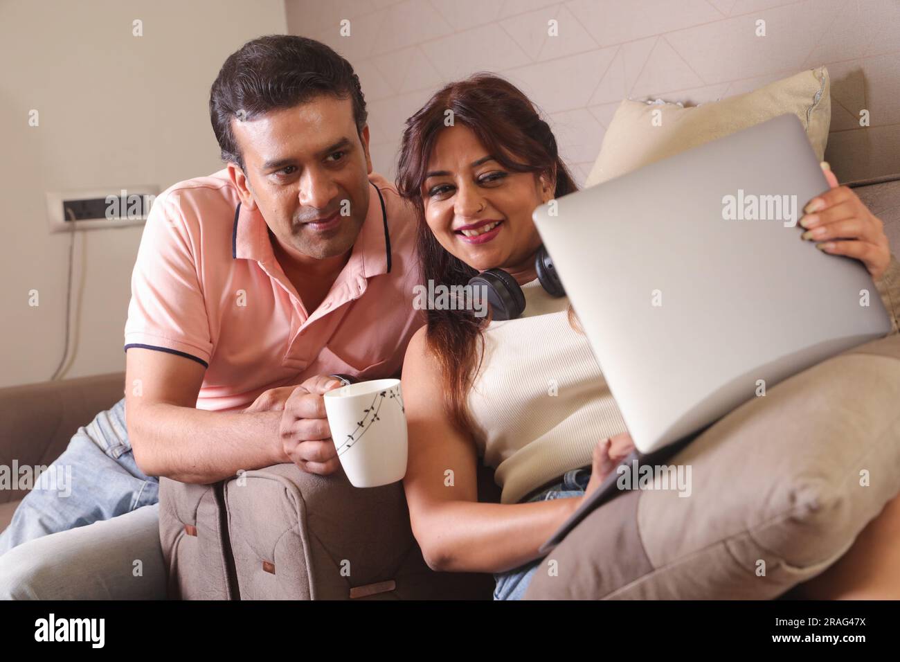Indisches glückliches Paar, das auf dem Sofa sitzt und mit einem Laptop online surft. Und eine Tasse Kaffee trinken und glückliche Momente. Stockfoto
