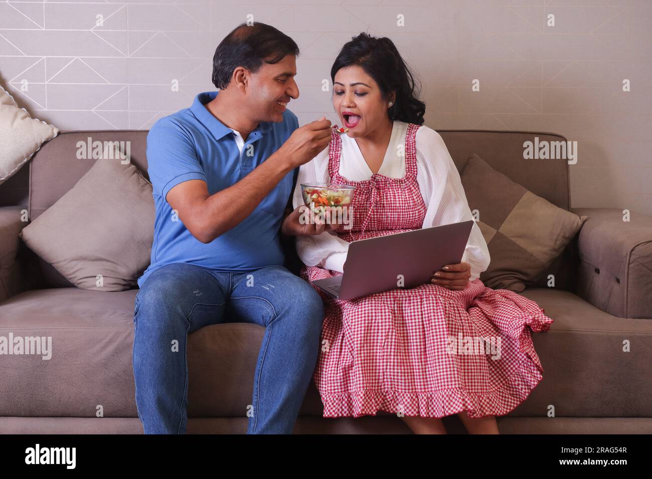 Indisches glückliches Paar, das Salat isst und eine gesunde Ernährung hat, auf einem Sofa sitzen und einen Laptop online surfen. Grüner Salat. Laptop in der Hand halten. Stockfoto