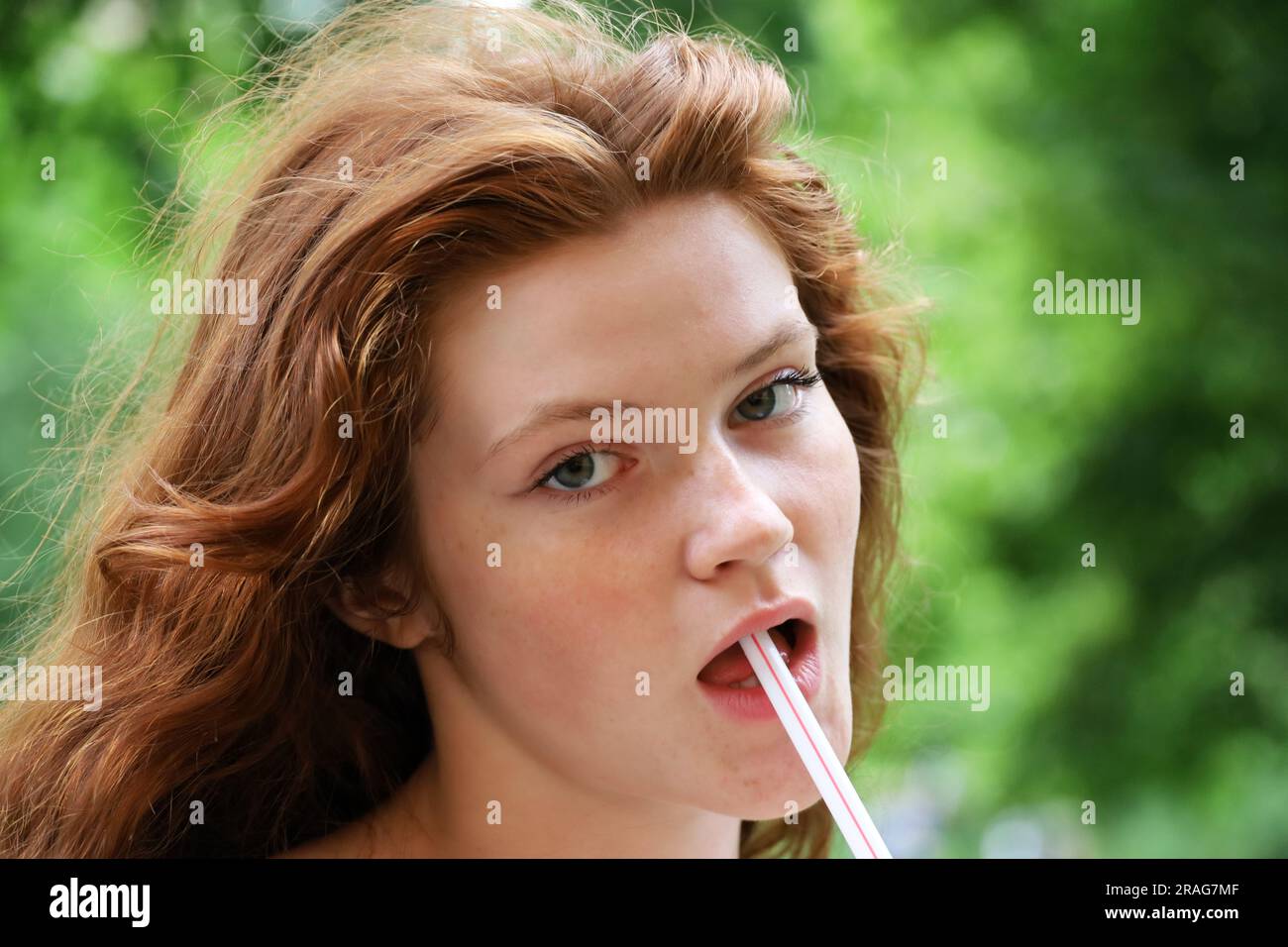 Portrait eines hübschen Ingwermädchens mit langen roten Haaren und Sommersprossen, das im Sommerpark einen Cocktail mit Strohhalm trinkt. Ein junges sinnliches Modell, weibliche Schönheit Stockfoto