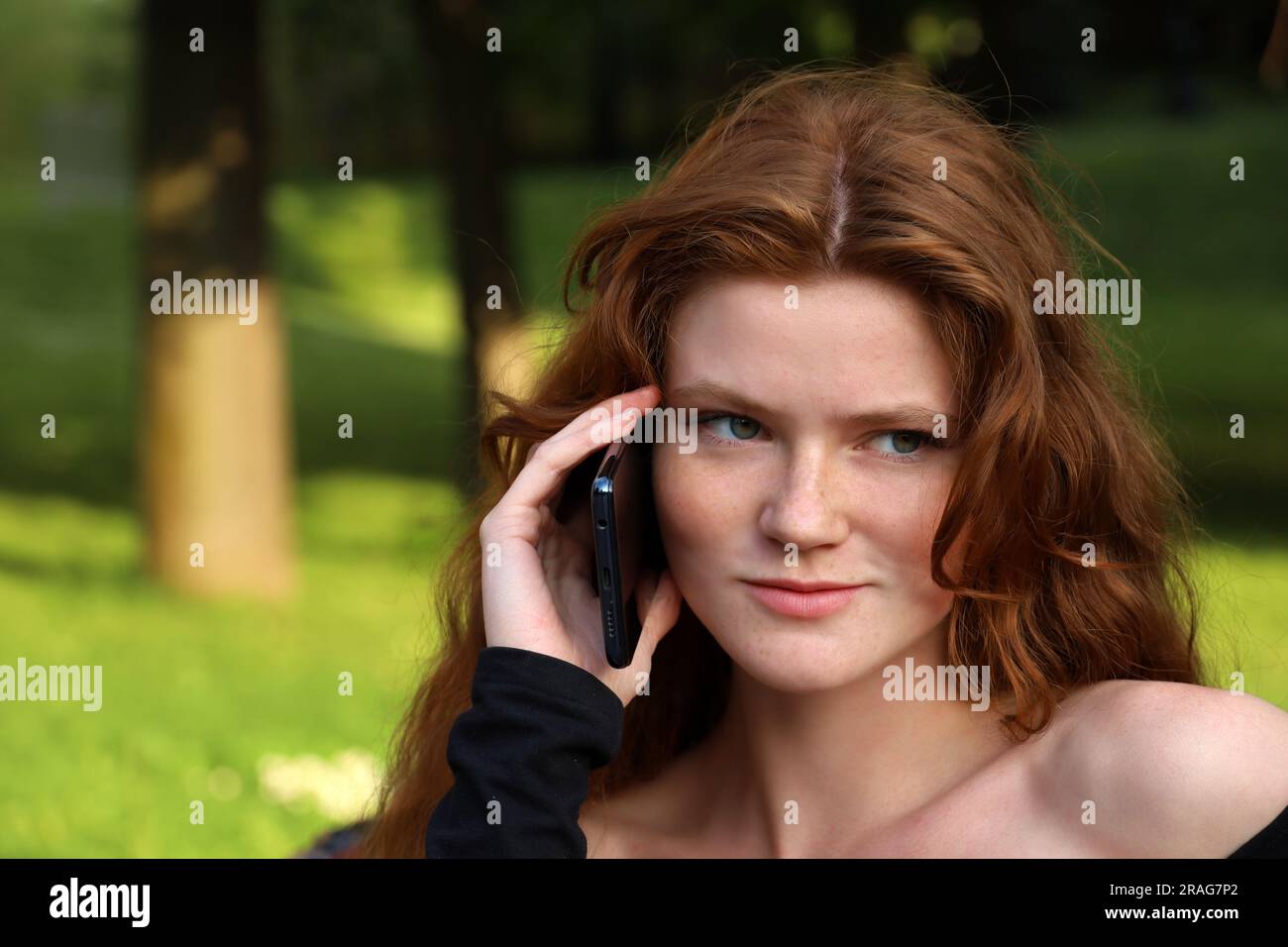 Lächelndes Mädchen mit langen Ingwerhaaren und Sommersprossen, das mit dem Handy telefoniert und auf einer Bank im Sommerpark sitzt Stockfoto