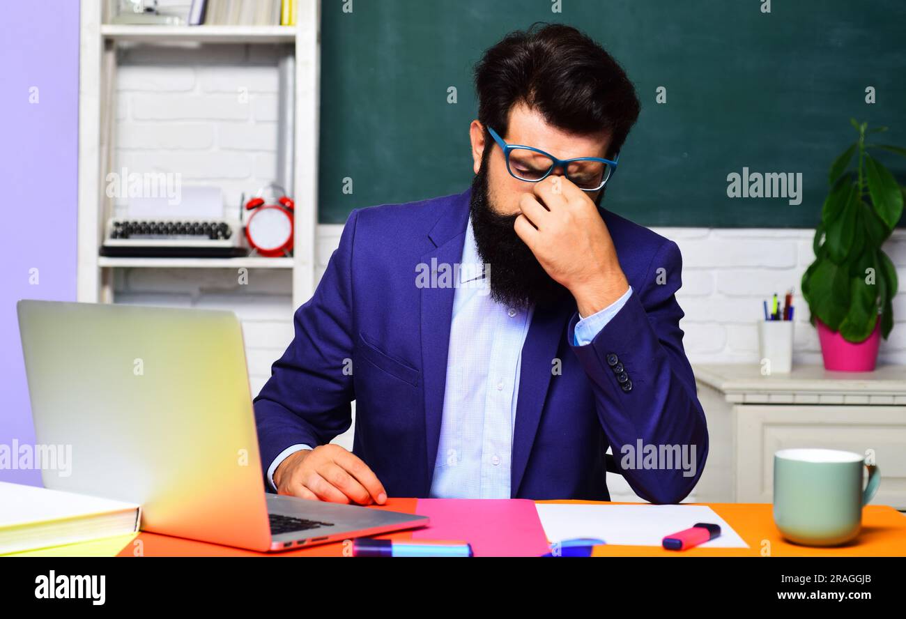 Bildung. Gestresster müder Schüler, der sich auf Tests oder Prüfungen im Klassenzimmer vorbereitet. Ermüdung nach langer Nutzung des Notebooks. Erschöpfter, bärtiger Lehrer fühlt sich verspannt Stockfoto