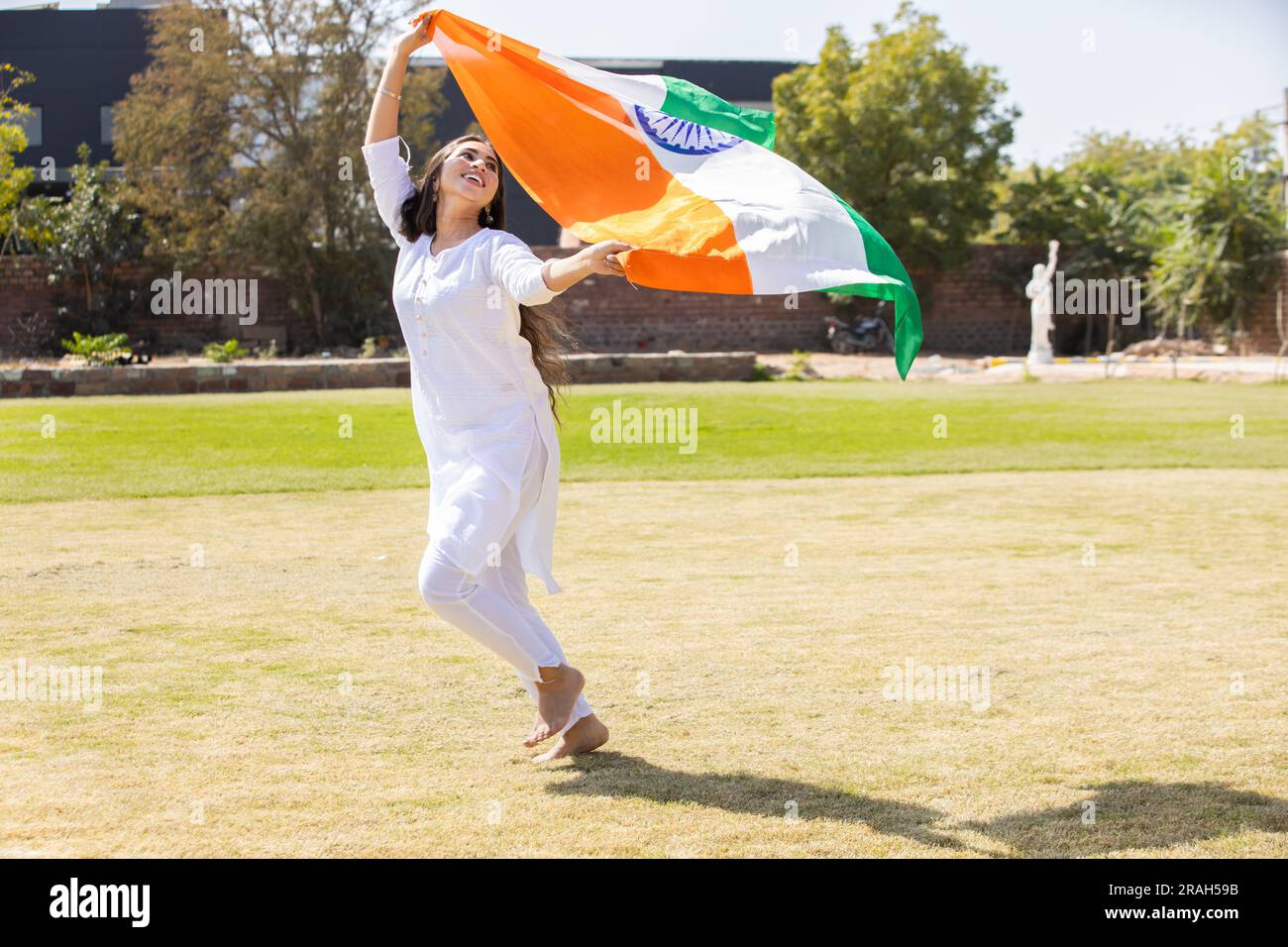 Glückliche junge Frau in traditionellem weißen Kleid, die im Park mit einer großen indischen Flagge läuft, um den Unabhängigkeitstag oder den Republikstag zu feiern. Stockfoto