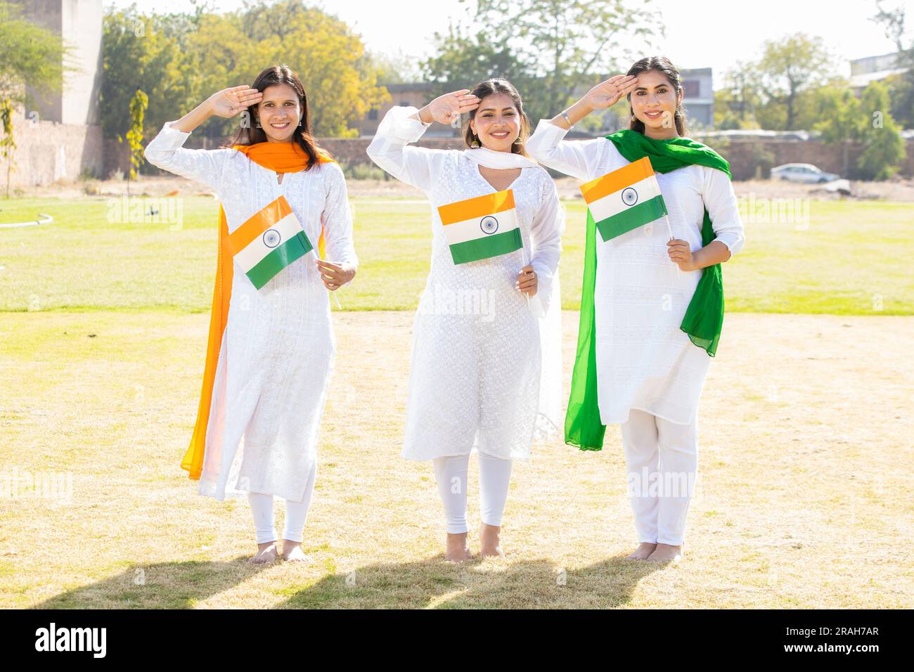 Drei junge, wunderschöne Frauen in traditionellem weißen Kleid mit indischer Flagge und Salut. Sie stehen im Park und feiern den Unabhängigkeitstag oder die Republik Stockfoto