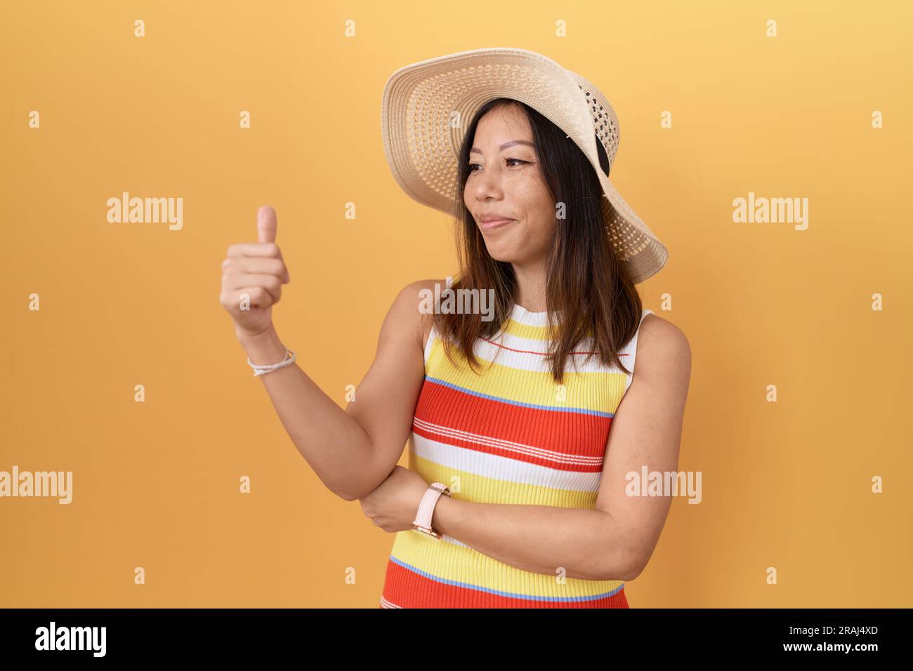mittelalterliche chinesin mit Sommerhut auf gelbem Hintergrund, die stolz aussieht und mit Daumen-hoch-Geste zur Seite lächelt Stockfoto