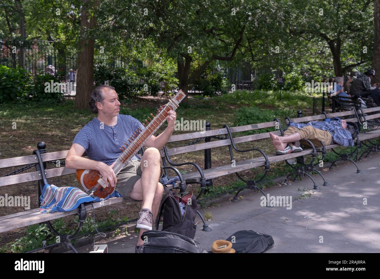 Eine New Yorker Straßenszene mit einem nicht-hinduistischen Sitarspieler in der Nähe eines schlafenden Schuhlosen. Im Washington Square Park in Greenwich Village, New York City. Stockfoto