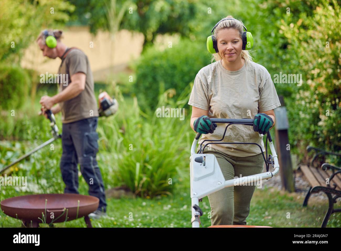 Junge Gärtnerin, die einen Rasenmäher verwendet, während ihr Kollege oder ihr Chef den Garten im Hintergrund trimmt Stockfoto